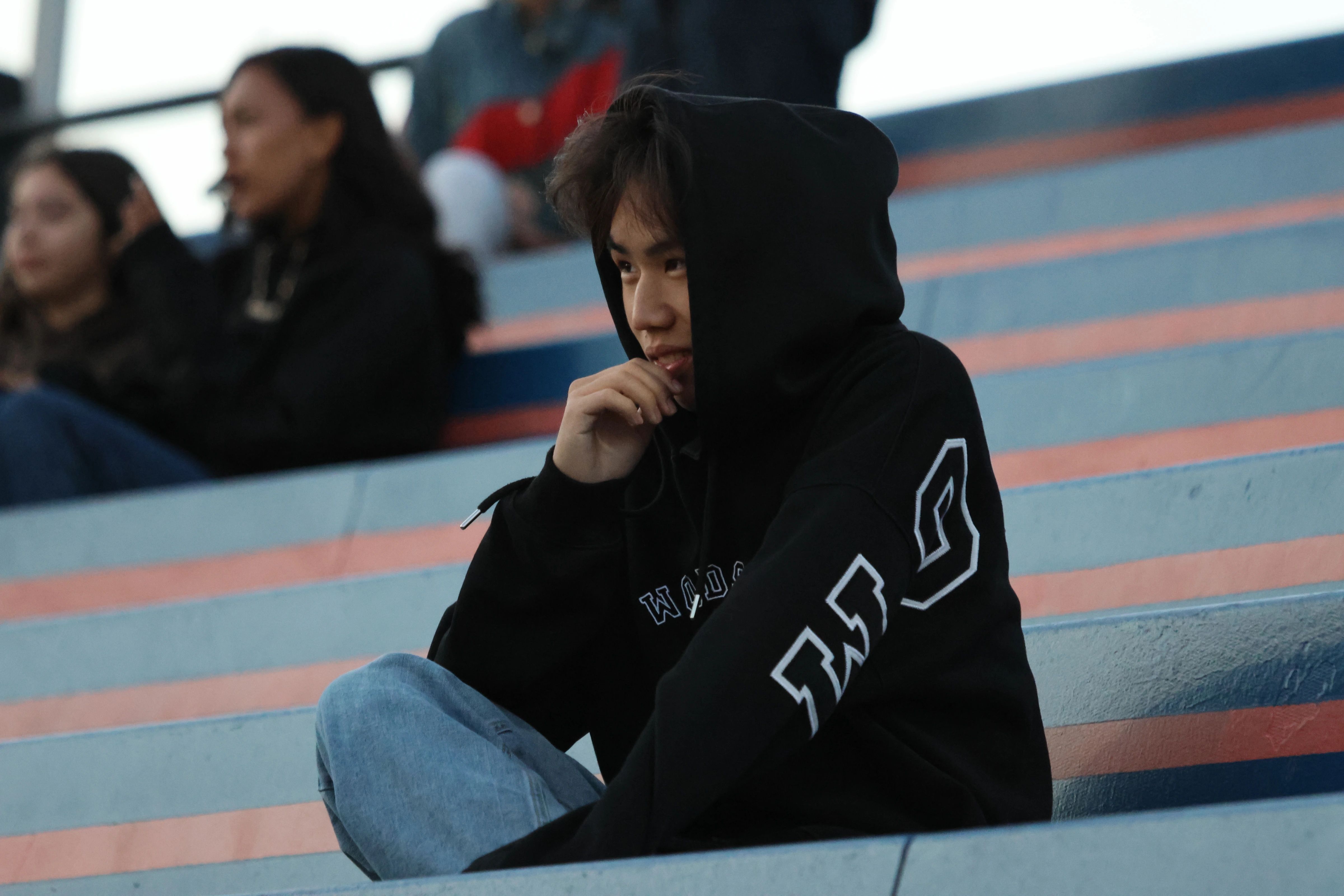 A young spectator in a black hood sits on striped stadium bleachers while looking sideways