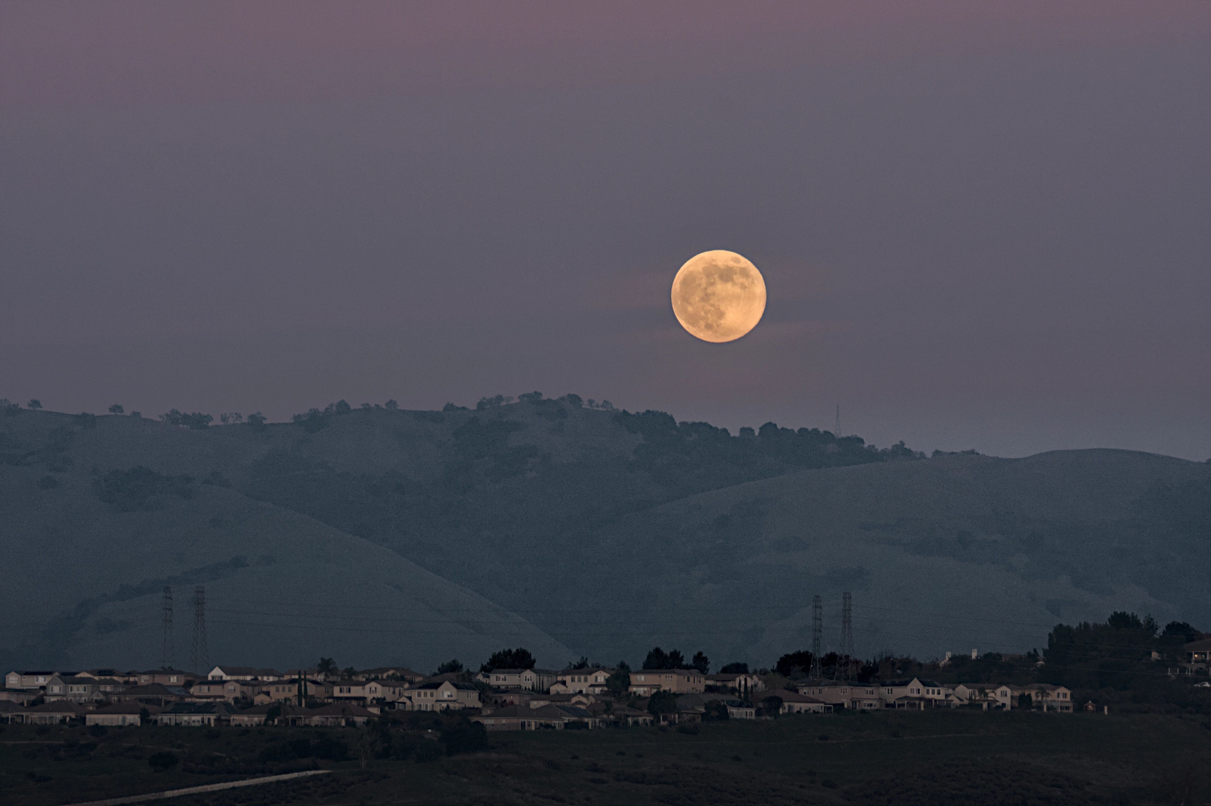 A full moon rising above dark rolling hills and a row of houses