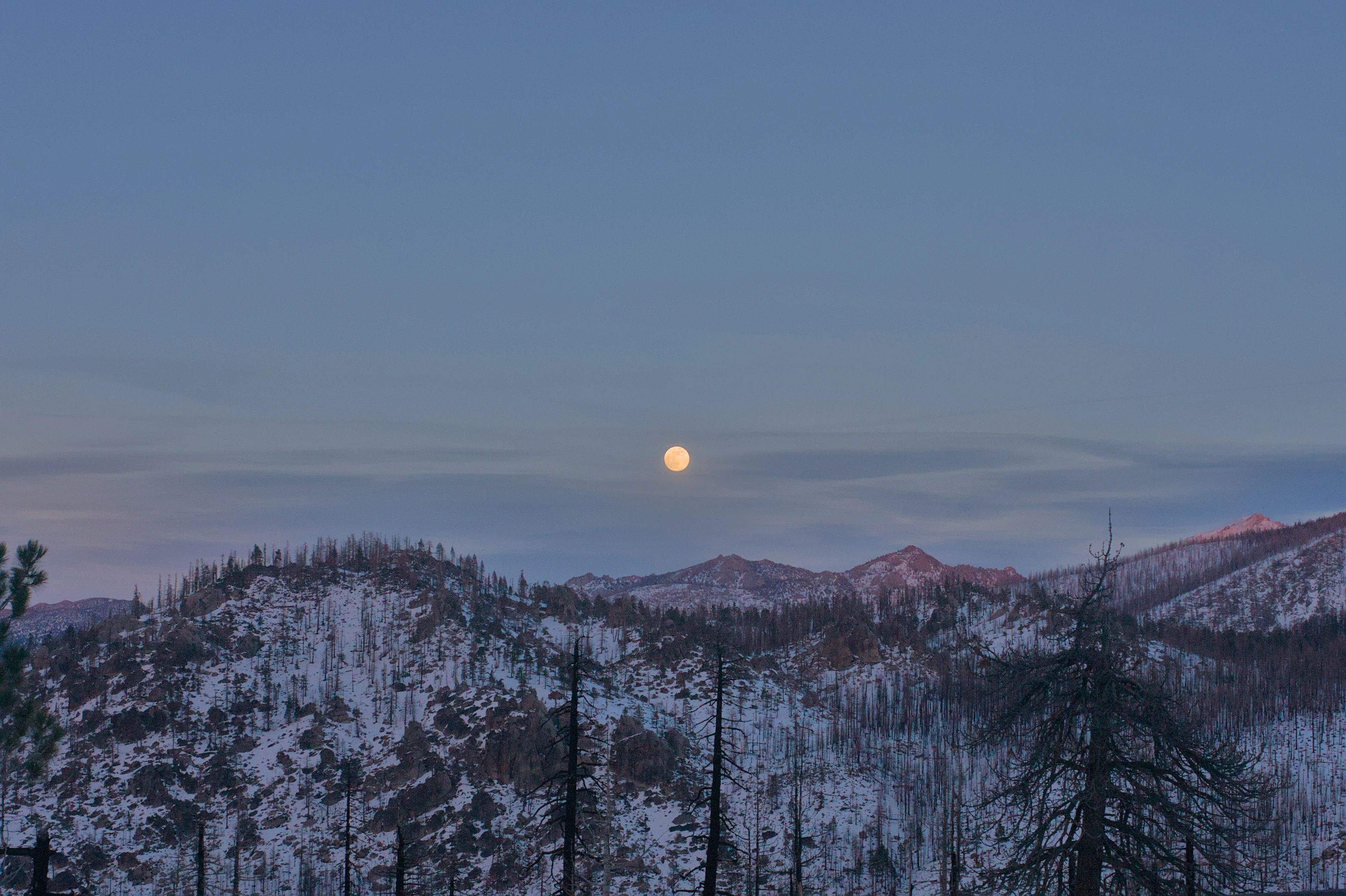 A pale moon suspended above a snow-covered mountain ridge at dusk