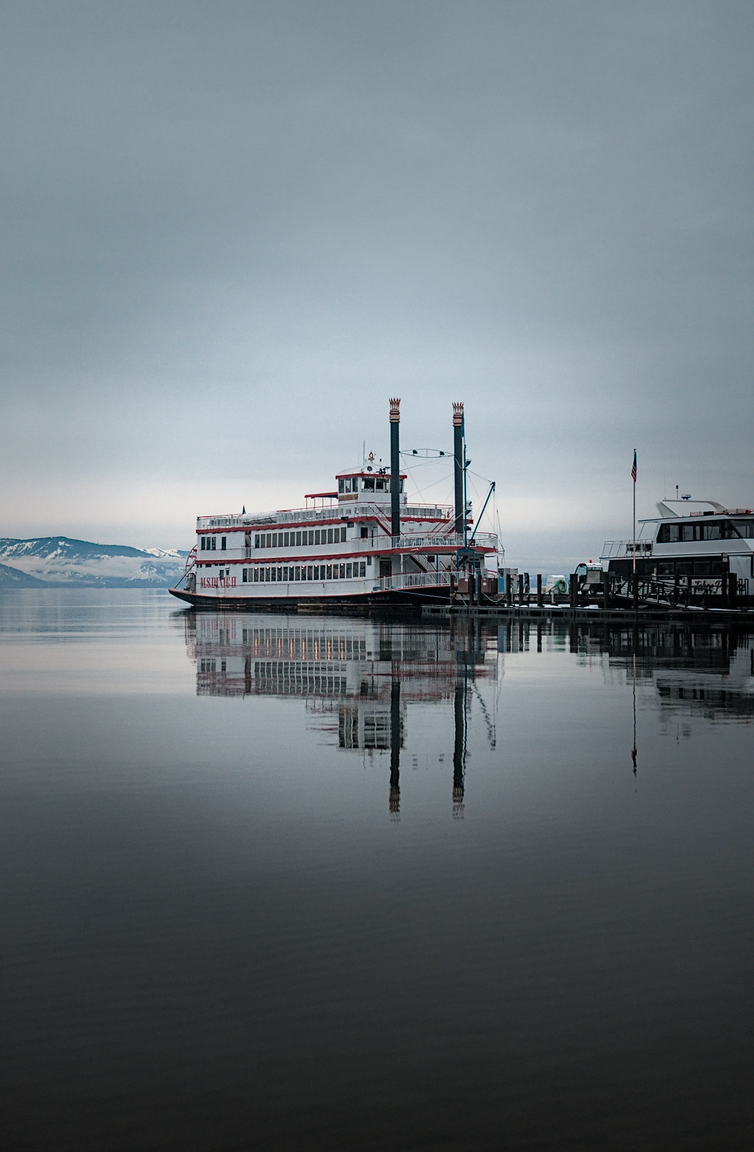 A paddlewheel boat and its reflection resting on the still gray water of Zephyr Cove