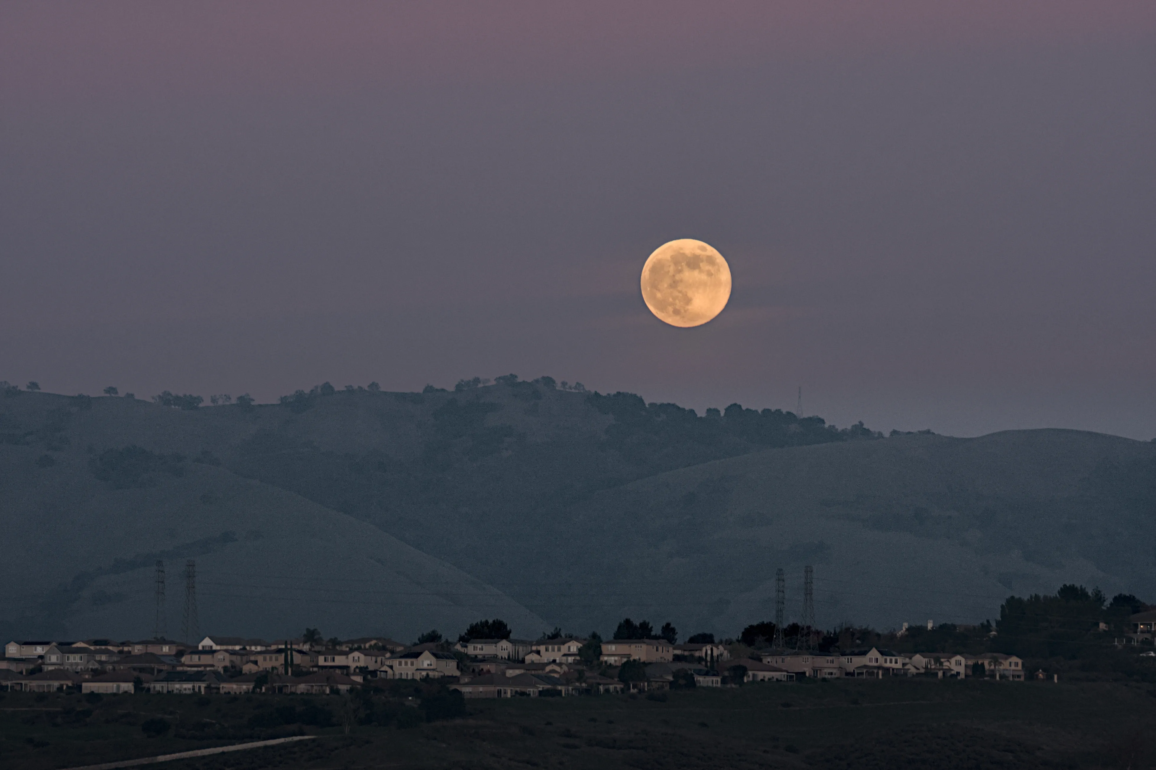 A full moon rising above dark rolling hills and a row of houses
