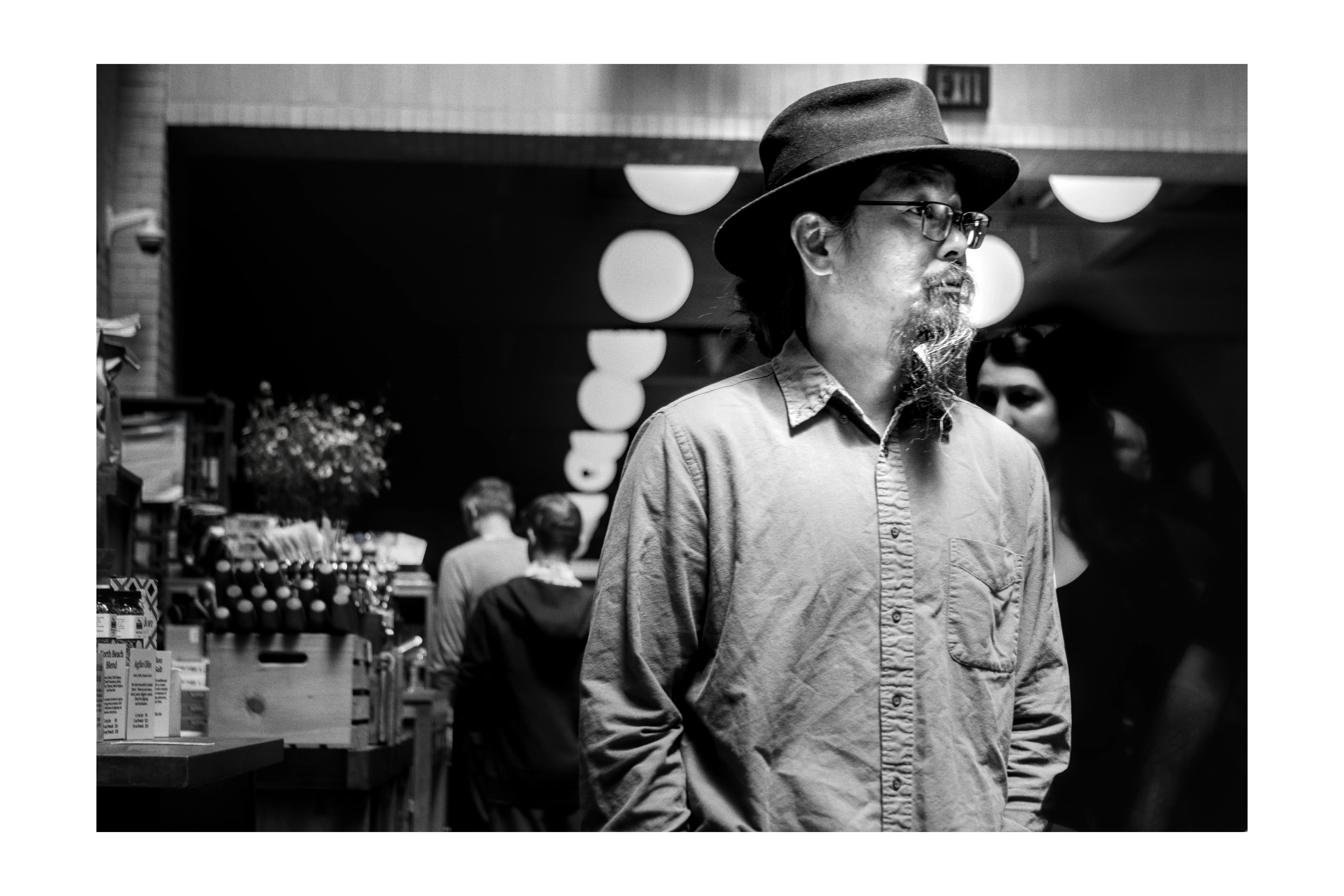 A man in a wide-brimmed hat and glasses passing beneath glowing market lights
