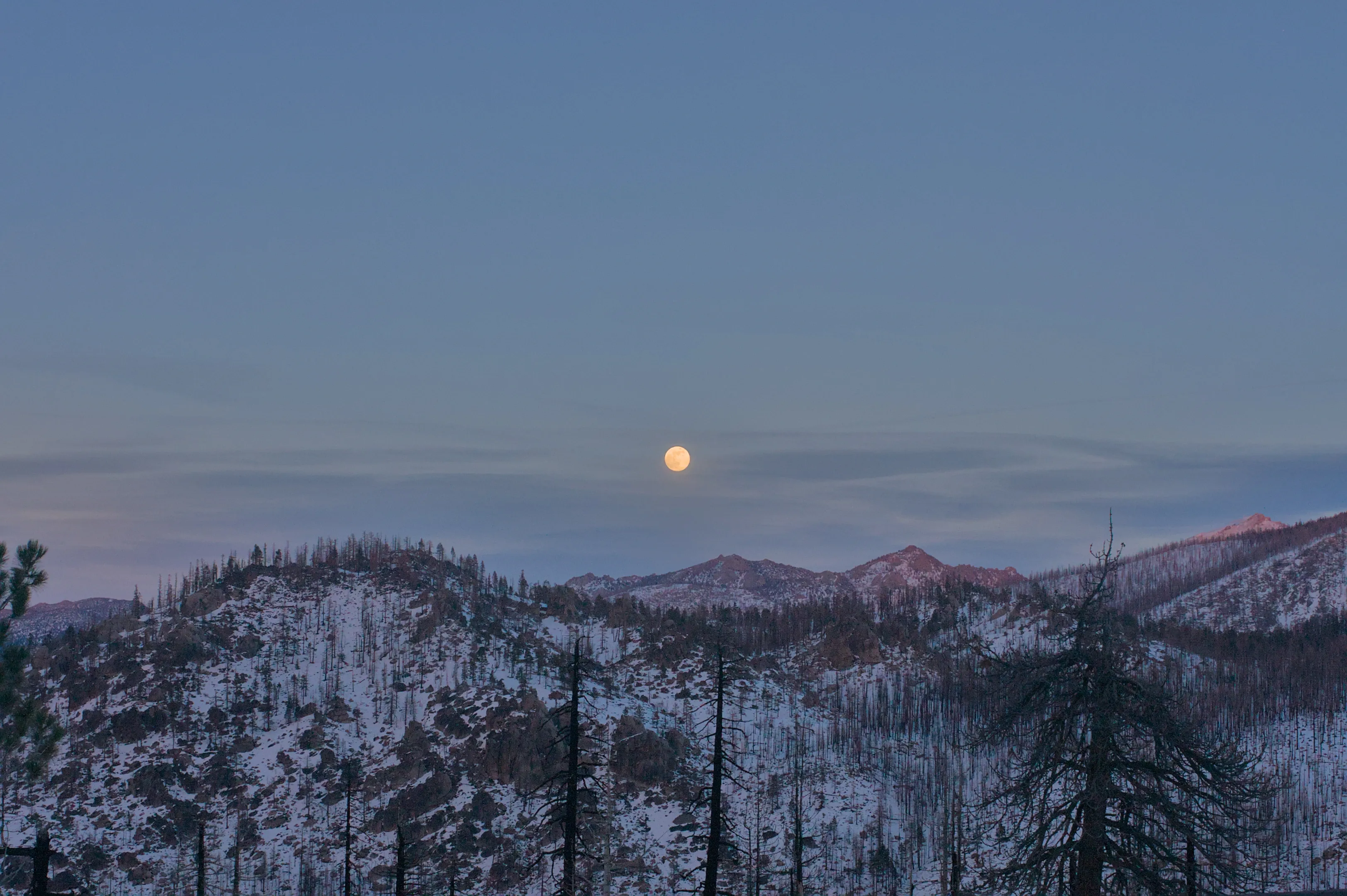 A pale moon suspended above a snow-covered mountain ridge at dusk